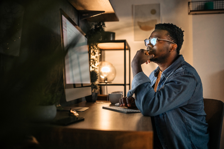 Young black man working late in the office, using desktop computer.